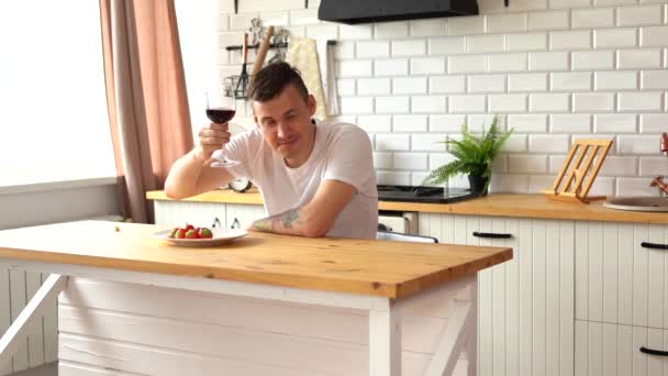Młody Szczęśliwy Człowiek Pijący Czerwone Wino Siedzący Przy Kuchennym Stole — Wideo stockowe