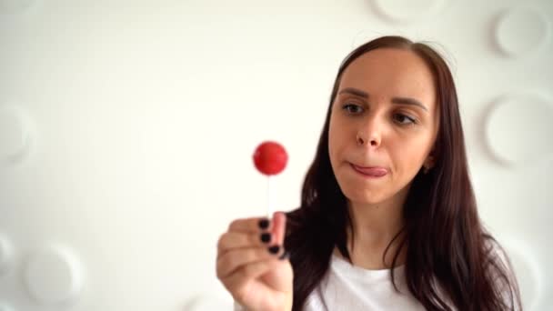 年轻女人在白色花纹的墙上舔大棒棒糖 接近成年的雌性用棍子吃糖果 — 图库视频影像