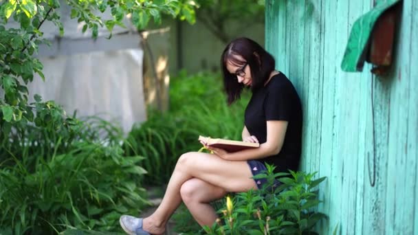 庭で本を読んでいる若い女性のクローズアップ 自然の中で休む女性 彼女の余暇を楽しんで — ストック動画