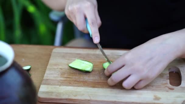 Dışarıdaki tahta tahtada yeşil salatalık kesen kadın. Kadın eli yakın çekimde doğrama tahtasındaki bıçakla sebzeleri kesiyor.. — Stok video