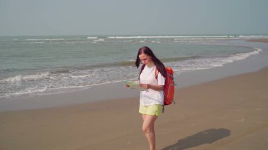 Sırt çantalı genç bir kadın deniz kıyısında yürüyor. Dişi turist dinlenirken harita okuyor ve inceliyor