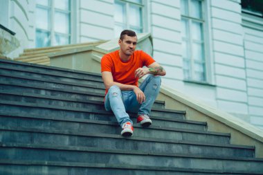 Kırmızı tişörtlü ve kot pantolonlu genç bir adam malikanenin merdivenlerinde poz veriyor. Kırmızı spor ayakkabılı yetişkin bir adam sarayın merdivenlerinde oturuyor..