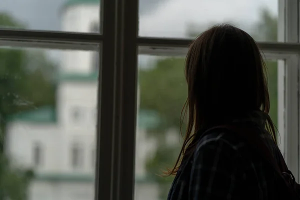 Close-up van een jonge vrouw die uit het raam kijkt in een onverlichte kamer. Vrouwelijke silhouet observeren door raam. — Stockfoto