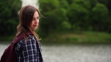 Gölün yanında duran genç bir kadın. Gölün ve evin bulanık arka planında dikilirken kareli gömlekli, gülümseyen ve başka tarafa bakan genç bir kadın.