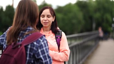 Sırt çantalı neşeli genç bir kadın. Üniversite eğitiminden sonra köprüde dururken gülümsüyor ve kız arkadaşıyla konuşuyor.