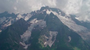 Bulutlu gökyüzüne karşı karlı dağ. Beyaz bulutların altından mavi gökyüzünde yüzen dağların üzerinden doğada güneşli bir günde karla kaplı.