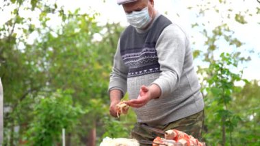 Tıbbi maskeli olgun bir adam şişe çiğ şiş saplar. Yaşlı erkek barbeküyle şiş tutuyor. Açık hava pikniği kavramı.