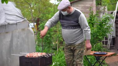 Tıbbi maskeli olgun bir adam ızgarada çapayla kömür karıştırıyor. Yaşlı bir erkek bahçede sulu barbekü yapıyor. Açık hava pikniği kavramı.
