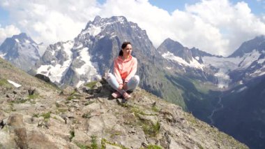 Genç bir kadın kayanın üzerinde oturuyor ve dağ manzarasına bakıyor. Dişi gezgin dağlık bölgede güzel manzaranın tadını çıkarıyor..
