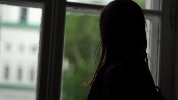 Κοντινό πλάνο μιας νεαρής γυναίκας που κοιτάει έξω από το παράθυρο σε ένα άφωτο δωμάτιο. Γυναικεία σιλουέτα παρατηρώντας μέσα από το παράθυρο. — Αρχείο Βίντεο