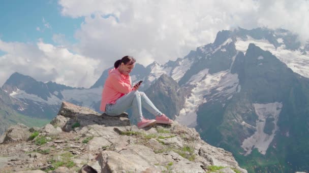 山の中でスマートフォンを使う女性旅行者 全身女は岩の上に座って 山の中で晴れた日に曇った空に対してスマートフォンを閲覧 — ストック動画