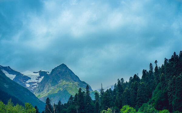 Beautiful mountain landscape in wooded area in summertime. Mighty mountains with snow and green array in cloudy weather.