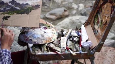 Dombay, Rusya 26 Temmuz 2020: Yürüyüş sırasında karton üzerine resim çizen genç bir kadın. Dışarıdaki dağ manzarasını resmeden bir ressam..