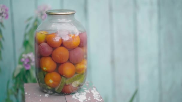 Grote glazen pot met gepekelde groenten op stoel op de achtergrond van oud houten huis. Tomaten en komkommers in blik in verzegelde pot op straat in de tuin. — Stockvideo