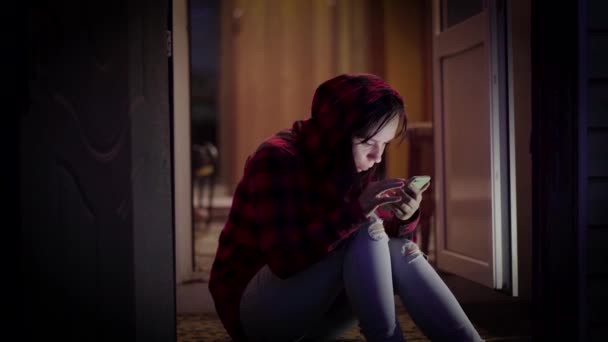 年轻妇女坐在门廊上 晚上浏览手机 成年女性在智能手机里翻来覆去地聊天 晚上坐在门廊上 — 图库视频影像