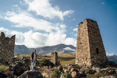 Dağlardaki eski harabelerin yanında duran bir gezgin. Yeşil tepede duran genç erkek turistin sisli ormanlı dağlara karşı yaşlanmış taş binalar ve görkemli manzaraya hayran olan manzarası..