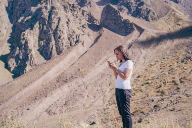 Genç bir kadın cep telefonuna bakıyor, tepede duruyor. Dişi gezgin yazın dağın arka planında akıllı telefonuyla durur