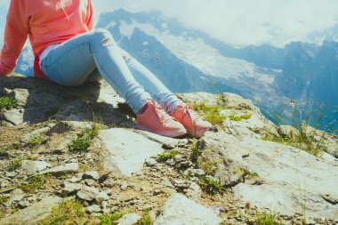 Güneşli bir havada kayanın üzerinde oturan genç bir kadın. Yazın dağın kenarında dinlenen kadın bir gezginin vücudunu kapatın.