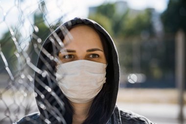 Kapüşonlu genç kadın portresi ve güneşli bir havada tıbbi maske. Yetişkin dişiye kafes yoluyla yaklaşın. Koronavirüs salgını sırasında kendinizi hastalıklardan koruyun.