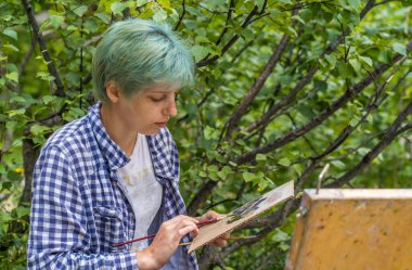 Genç bir kadın palette boya karıştırıyor, yürüyüş yaparken karton üzerine resim çiziyor. Ressam renkleri karıştırır, dağ manzarası çizer..