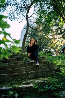 Yeşil botanik parkında yürürken eski taş merdivenlerde oturan ve kahve içen genç bayan gezginin alçak açısı..