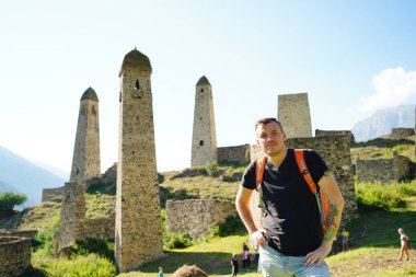 Sırt çantalı genç adam muazzam dağların arkasındaki eski kulelerin yanında duruyor. Erkek turist güzel manzaranın tadını çıkarıyor, dağlık bölgedeki yıkık tarihi binalarda duruyor.