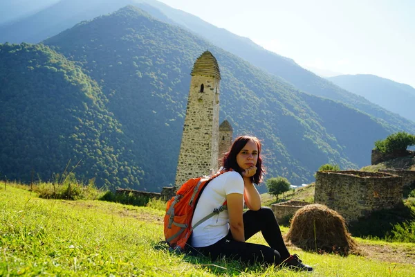 Carefree traveling woman relaxing on green lawn. Side view of serene female tourist with backpack sitting on green hill and enjoying sunny weather on background of mountainous landscape.