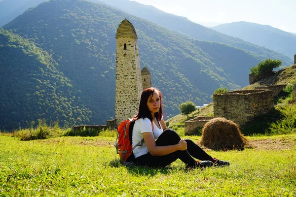 Carefree traveling woman relaxing on green lawn. Side view of serene female tourist with backpack sitting on green hill and enjoying sunny weather on background of mountainous landscape.