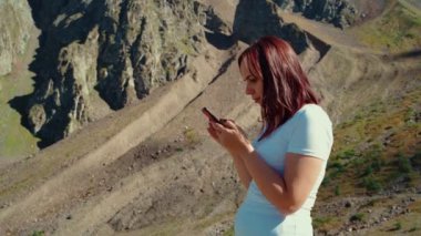 Genç bir kadın cep telefonuna bakıyor, tepede duruyor. Dişi gezgin yazın dağın arka planında akıllı telefonuyla durur