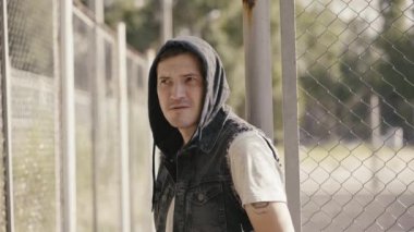 Kapüşonlu genç adam, çitin yanında dururken dudaklarını ısırıyor. 