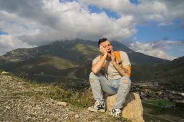 Yorgun bir seyyah tepede dinleniyor. Yorgun bir erkek turist taşın üzerinde oturuyor ve dağlarda aktif bir yürüyüş yaptıktan sonra tatlı tatlı esniyor..