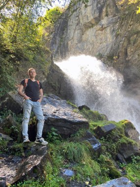 Şelalenin arka planına bakan genç adamın portresi. Yetişkin erkek turist dağlık bölgede güzel manzaranın tadını çıkarıyor..
