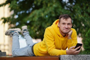 Genç adam dışarıda bir bankta uzanmış, bacakları yukarda ve cep telefonunu karıştırıyor. Akıllı telefon kullanan bir erkek, konut binasının avlusunda internet kullanıyor.