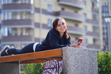 Genç bir kadın, dışarıda bir bankta uzanmış, bacakları yukarda ve cep telefonunu karıştırıyor. Akıllı telefon kullanan bir kadın bir apartmanın avlusunda internet kullanıyor..