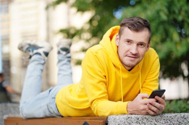 Genç adam dışarıda bir bankta uzanmış, bacakları yukarda ve cep telefonunu karıştırıyor. Akıllı telefon kullanan bir erkek, konut binasının avlusunda internet kullanıyor.