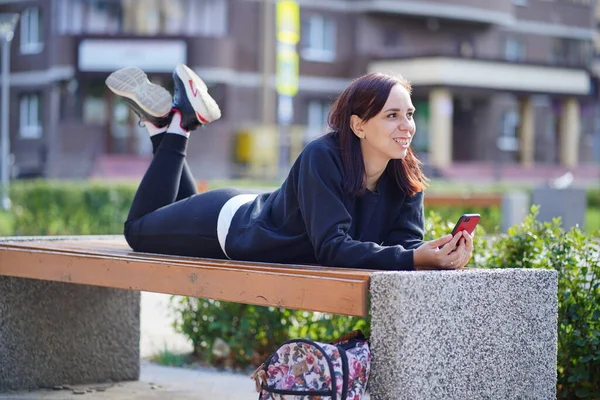 벤치에 다리를 여성이 핸드폰을 있습니다 스마트폰을 사용하는 여자가 뜰에서 인터넷을 — 스톡 사진