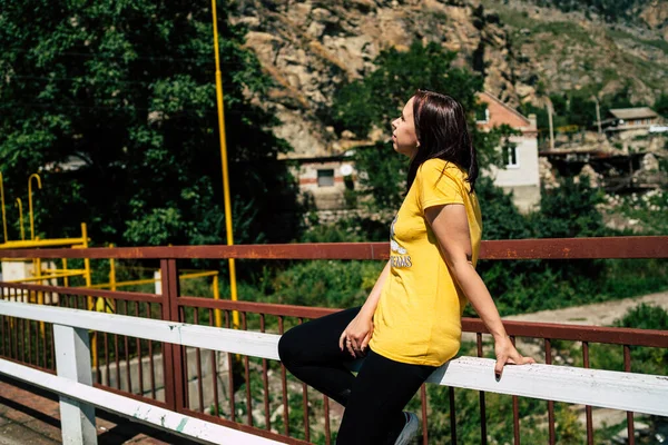 Tanınmayan genç kadın köprüde duruyor ve yeşil ağaçlar arasında akan dağ nehrinin güzel manzarasının tadını çıkarıyor. Kadın turistin temiz su yoluna baktığı arka manzara.