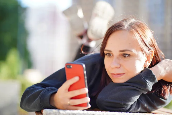 Genç bir kadın, dışarıda bir bankta uzanmış, bacakları yukarda ve cep telefonunu karıştırıyor. Akıllı telefon kullanan bir kadın bir apartmanın avlusunda internet kullanıyor.