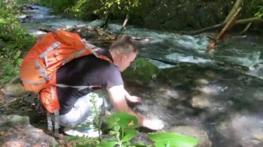 Sırt çantalı genç adam dağ nehrinin yanında çömelip içiyor. Erkek turist, dağlarda aktif bir yürüyüş yaptıktan sonra susuzluğunu gidermek için temiz suyu elleriyle temizler.