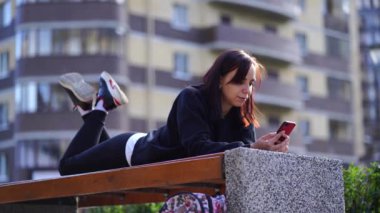 Genç bir kadın, dışarıda bir bankta uzanmış, bacakları yukarda ve cep telefonunu karıştırıyor. Akıllı telefon kullanan bir kadın bir apartmanın avlusunda internet kullanıyor..