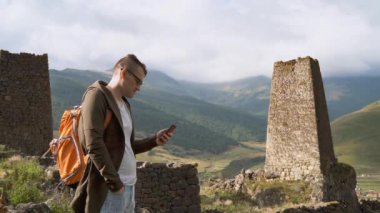 Sırt çantalı ve akıllı telefonlu genç adam muazzam dağların arkasındaki eski kulelerin yanında duruyor. Erkek turist cep telefonu, dağlık bölgedeki yıkık tarihi binalarda duruyor.