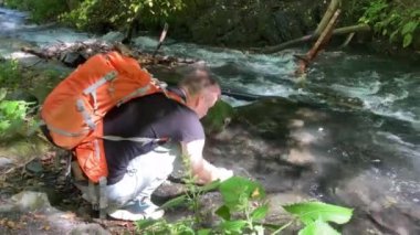 Erkek gezgin yüzünü dağ suyuyla yıkar. Dağ nehrinin kıyısındaki kayalıklarda oturan turist kendini tazeliyor..