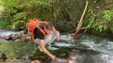 Sırt çantalı genç adam dağ nehrinin yanında çömelip içiyor. Erkek turist, dağlarda aktif bir yürüyüş yaptıktan sonra susuzluğunu gidermek için temiz suyu elleriyle temizler.