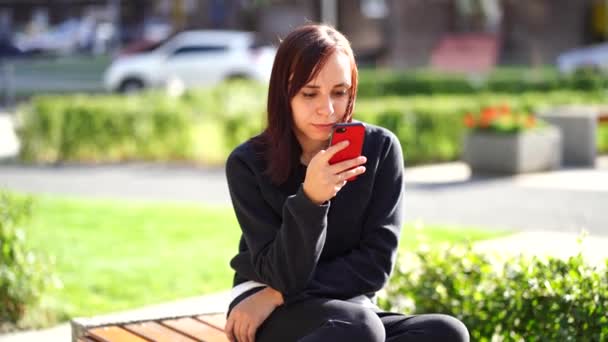 公園のベンチに座っている若い女性は 携帯電話を見ている スマートフォンを使っている女性が住宅の中庭でインターネットを利用しています — ストック動画