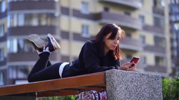 若い女性は彼女の足でベンチの外の彼女の胃の上に横たわって 彼女の携帯電話を参照してください スマートフォンを使っている女性が住宅の中庭でインターネットを利用しています — ストック動画