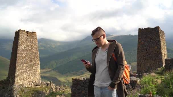 バックパックとスマートフォンを持つ若い男は、強大な山々を背景に古い塔の近くに立っています。山間部の破壊された歴史的建造物に立つ男性観光客の携帯電話を閲覧 — ストック動画
