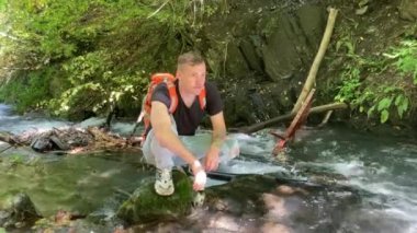 Sırt çantalı genç adam dağ nehrinde taşın üzerinde oturur ve yüzünü çevirir. Erkek turist dinleniyor ve yaz aylarında güzel bir çevre manzarası izliyor.