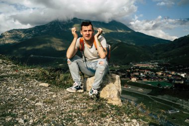Genç adam, dağlardaki köyün arka planında taşın üzerinde oturuyor. Yorgun erkek turist, dağlarda aktif bir yürüyüşten sonra tepede dinleniyor.