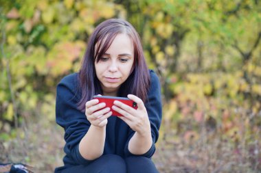 Günlük giysiler içinde seyir defterinde oturan ve cep telefonunu karıştıran genç bir kadın. Yetişkin esmer ormanda akıllı telefonuyla dinleniyor..