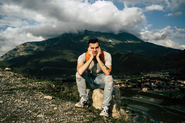 Young man grimaces, sitting on stone on background of village in mountains. Tired male tourist aping, resting on hill after active trekking in mountains